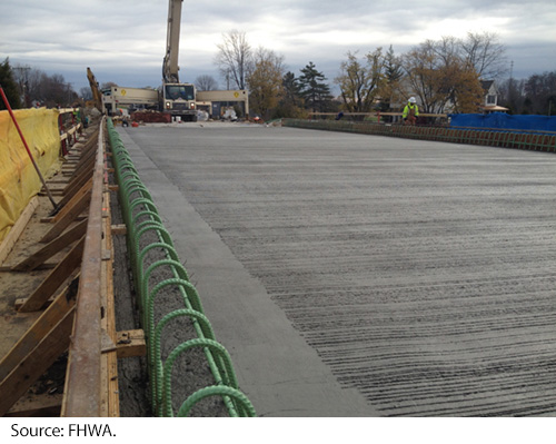 Uncured concrete on a reinforcing-rod-lined roadway with a crane and a worker in the distance. Image Source: FHWA.