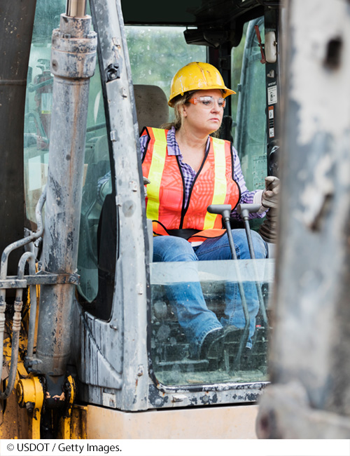 A female highway worker wearing a safety vest and googles operates a tractor. Image Source: © USDOT / Getty Images.