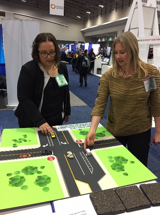 Two people use toy cars on a tabletop demo of dedicated left- and right-turn lanes.