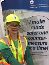 A woman in a hard had and retroreflective vest stands in front of a 'Selfie Station' with a bubble on a banner that reads 'I make roads safer one countermeasure at a time.'