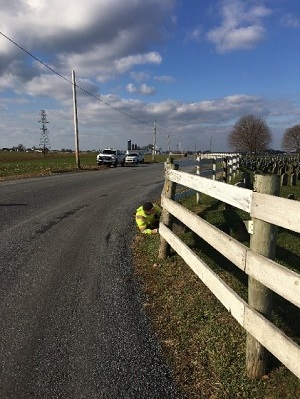 Photo shows a person working on a fence along a road curve with very little to no shoulder space.