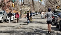 Photo shows cars parked along a street with people walking and biking.