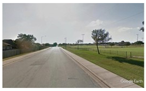 Photo depicting the approach to a 90-degree curve from a distance. A pedestrian sidewalk runs along the right side of the roadway and extends straight into the distance beyond the sharp left curve of the roadway. At this distance, it appears that the roadway and the pedestrian path both extend straight ahead.