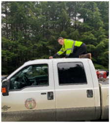 This photo shows a woman wearing retroreflective gear on top of the cab of a pickup truck attaching a camera to the roof.