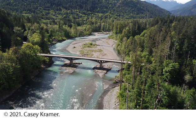 "Drone photo looking down at an angle on the U.S. 101 bridge spanning the Elwha River in a heavily forested location. Rolling mountains are seen in the background. The flowing river water bends around a large point bar of sediment and then flows through the three spans of the bridge. Image Source: © 2021. Casey Kramer."