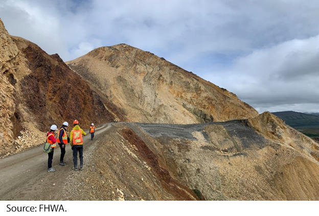 "Four field crew members, dressed in safety vests, stand at the edge of the landslide along a gravel road. Image Source: FHWA."