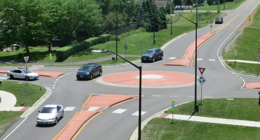 "A single-lane roundabout where each of the four legs approaching the roundabout is a yield-controlled, two-lane roadway. Each approach leg features a high visibility crosswalk. Raised medians are present in the vicinity of the roundabout but not beyond the extents of the roundabout. The roundabout island appears to be traversable by trucks and other large vehicles. 