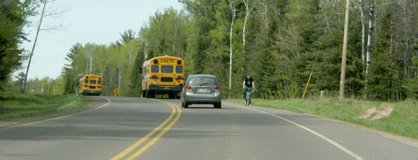 "Photo.  A two lane rural road with a shoulder only on the right side.  A cyclist is traveling against traffic on the shoulder.  "