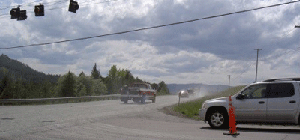 Photo of the westbound view of US Highway 2 from Springcreek Road. There are signal heads in the top left corner of the photo and a vehicle stopped at the intersection with a traffic cone in fluorescent orange shown in front of it. 3 vehicles are driving away in the image. A guardrail is seen on the left side with trees and mountains in the background.