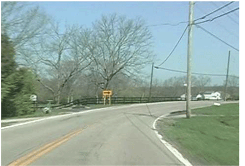 alt="Photograph of a horizontal curve on a rural, two-lane road before signage updates. The curve has centerlines and edge lines, but only has one one-direction large arrow sign within the curve."