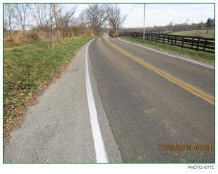 A two-lane rural roadway with darker pavement in the travel lanes indicating that they have been treated with HFST. Photo: KYTC