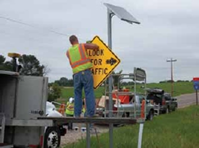 "Photograph of a worker on a truck affixing a yellow caution sign ('Look For Traffic') to a pole"