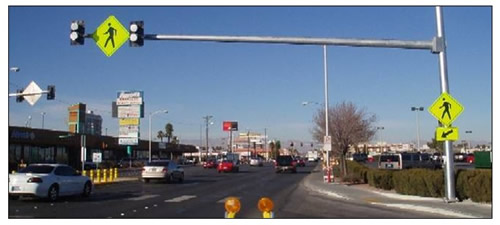 Photo of flashing beacons and a large pedestrian warning sign mounted to a mastarm suspended above a crosswalk at a busy intersection at a shopping area.