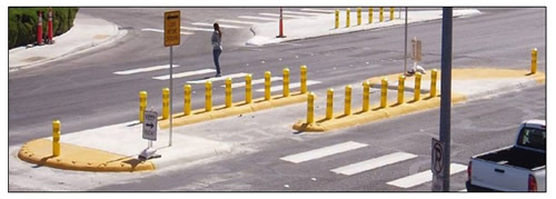 Photo of a refuge island in the middle of a multilane crossing characterized by bollards running parallel to traffic along the edges of the island with an opening on each side for pedestrians to pass, offset crosswalks on either side of the island structure, and an open center cutout area that channels pedestrians into a path that is parallel to the traffic for a few feet before turning and allowing pedestrians to traverse the second half of the crosswalk.