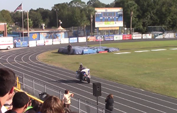 "Screen capture from the safety video shows a police officer riding a motorcyle on the track around a high school field while students watch from the stands"