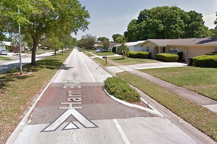 ""Figure 3.12.2. Speed Table with Choker. This figure contains a photograph of a residential area. On the left hand side of the photo there is a lane of trees on a landscaped median. To the right of the trees there is single lane road labeled Harn Blvd. A speed table, dyed a dusty pink, fills most of the lane. This traffic feature also uses a choker, a crescent shaped curb extension landscaped with shrubs that extends onto the speed table. To the right of that, there is a sidewalk separating the street from a row of single story houses.