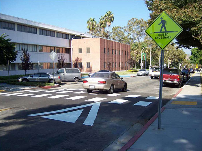 "Figure 3.14.2. Typical Raised Crosswalk Application. This figure contains a photograph of a two lane street in a business district. A sidewalk on the right hand side of the picture has a signpost holding a diamond shaped yellow sign showing an icon of a person walking over the words Raised Crosswalk. Between the curb and the right lane there is parallel parking where a red truck and another vehicle are parked. A light colored car and another truck have just crossed the walk in the right hand lane. The left lane is free of traffic. A raised crosswalk connects the sidewalks on the sides of the street."