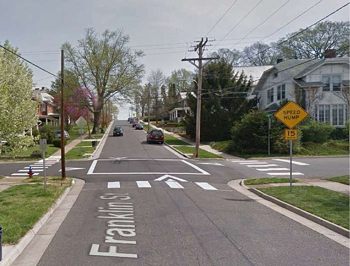 "Figure 3.15.3. Raised Intersection in Residential Setting without Raised Crosswalks. This figure contains a photograph of undivided two lane roads intersecting at the center of the photo. The road running from top to bottom, Franklin St., features parallel parking on the right hand side and a car approaches the intersection. There is a signpost to the right of Franklin, at the intersection, with a yellow diamond sign with the words Speed Hump and a square yellow sign below that which contains the text 15 MPH. There are houses on the opposite side of the intersection. Arrows painted between the crosswalks and the raised pavement indicate right of way. The raised intersection is bordered in white stripes and sidewalks."