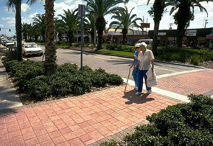 "Figure 3.17.3. Choker in Urban Commercial Setting. This figure contains a photograph of a two lane street. The street runs from top left to bottom right of the picture. Palm trees line each side of the street. A crosswalk runs across the bottom of the picture. A choker has been used to narrow the street at the crosswalk. Both crosswalk and sidewalk are made of patterned brick. Two older pedestrians have just crossed the street from right to left."