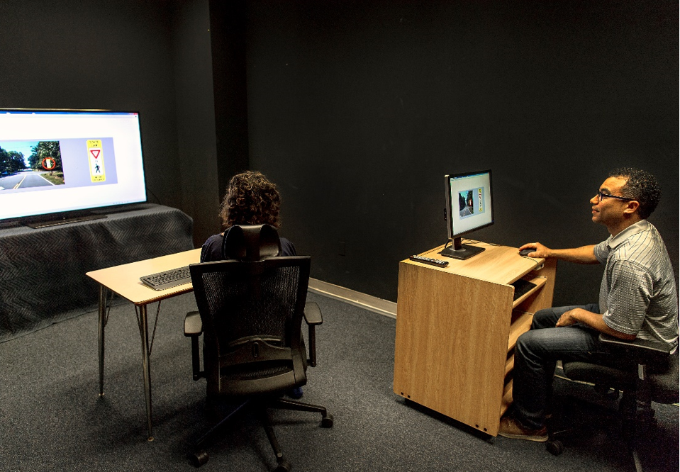 "A volunteer, seated at a desk with a keyboard, looking at a monitor. A researcher sits behind her, at his own desk with a computer.""