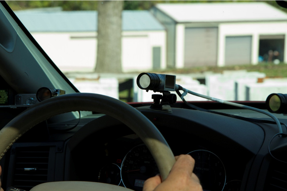 "A close up of a car dashboard, showing a volunteer's hands on the steering wheel, and beyond that a eye-tracking device mounted on the dashboard, facing the volunteer's eyes."