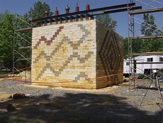 Figure 18. This photo shows a GRS abutment experiment built with a concrete masonry units (CMU) facing element on the outdoor strong floor. The CMU blocks have a zigzag pattern in different colors.  A concrete footing is positioned on top near the edge of the face of the GRS abutment with five hollow core hydraulic jacks on top. The hydraulic jacks are connected to load cells. Scaffolding flanks both sides of the GRS abutment experiment and reference beams are supported on the scaffolding to measure vertical deformations.