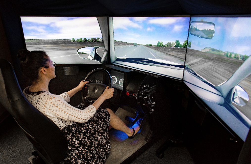 "A volunteer seated in the mock driver seat, hands on the steering wheel, navigating a simulated environment shown on multiple screens designed to simulate windows of a car."