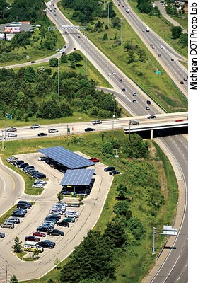 Photo. Vehicles are parked under a solar carport on a sunny day. Traffic passes by on adjacent highways.