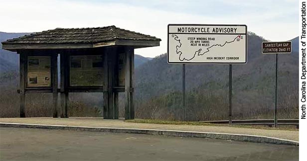 Photo. A scenic pulloff on a highway includes a large white sign that features an outline of the winding road ahead and a motorcycle advisory. The text reads, “Steep winding road, 20 mile per hour turns, next 18 miles, High Incident Corridor.