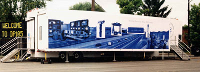 A parked white trailer that is painted with a blue mural of a city view, with trees in background of trailer