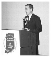 J. Bryan Nichol, wearing a suit while standing behind podium giving a speech