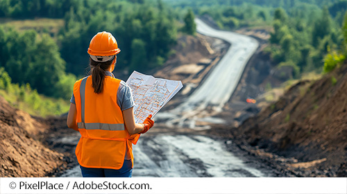 A woman wearing a hardhat and safety vest holds construction plans and looks out on a road made of dirt surrounded by trees. Image source: © PixelPlace / AdobeStock.com.