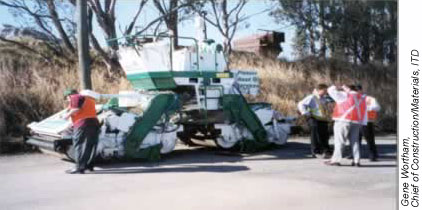 Three men standing near an asphalt compactor with trees and weeds in the background