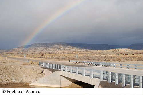 A roadway and bridge intersects a mountainous area with a rainbow in the sky. Image Source: © Pueblo of Acoma.