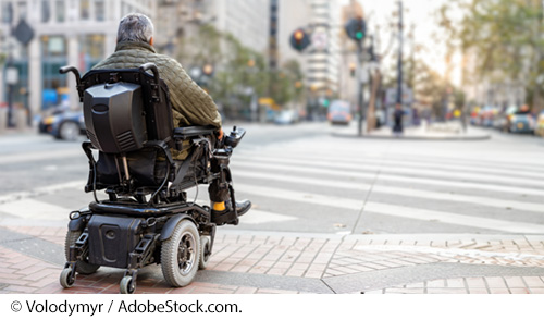 A man in a motorized wheelchair at a crosswalk, waiting to cross an urban street. Image © Volodymyr / AdobeStock.com.