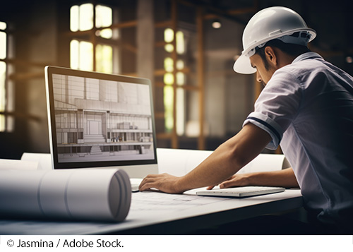 An engineer working on a blueprint in a digital design program on the computer. The work is performed in an office in a warehouse with scaffolding and viewed on a large desktop screen next to a roll of schematics. Image Source: © Jasmina / Adobe Stock.