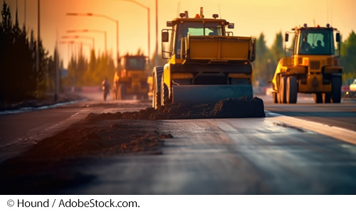 Three pavement construction vehicles stop on an asphalt pavement. A construction worker is standing near the construction vehicles. Image © Hound / AdobeStock.com.