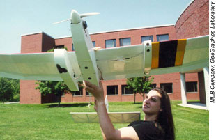 A woman  wearing a black shirt, holding a model unmanned aerial vehicle with a building and grass in the background