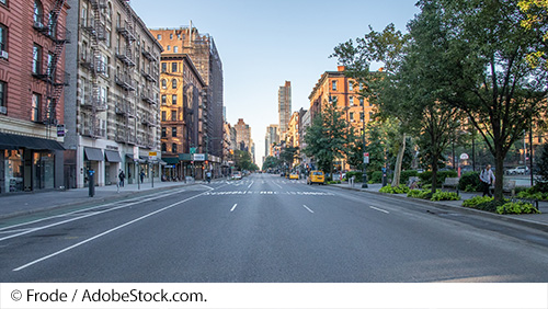 Street level view of an urban street with roadway markings for three traffic lanes and one separate space allocated for bicycles. Apartment buildings on the left side of the road and medium density of trees in the median line the right side of the road. Image Source: © Frode / AdobeStock.com.