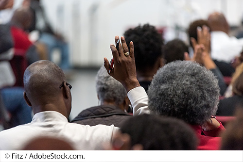 People raise their hands to ask questions during a crowded community meeting. Image Source: © Fitz / AdobeStock.com.