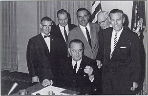 Administrator Rex Whitton (right) receives one of the pens that President Lyndon B. Johnson used to sign the Federal-Aid Highway Act of 1964. On Mr. Whitton's right is Commerce Secretary Luther Hodges, on his right is an unidentified gentleman (center), Representative Ed Edmundson (OK) and Representative Frank M. Clark (PA)-both members of the Committee on Public Works. Also at the ceremony were members of the Cabinet, the Congress, and representatives of the highway industry.