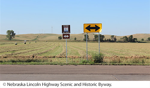 A view of a roadway marked with a “Historic 1913 Route” sign. Along the road is a grassy field scattered with haybales, trees, and powerlines atop hills in the distance. Image Source: © Nebraska Lincoln Highway Scenic and Historic Byway.