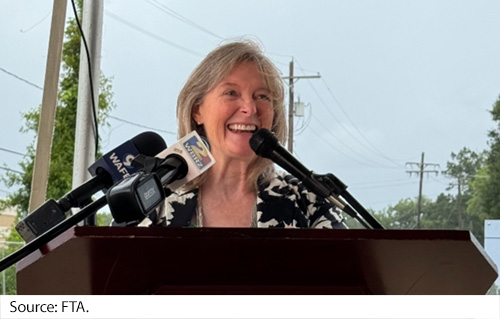 A female stands at a podium with microphones extended toward her face. Image Source: FTA.