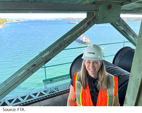 A female wearing a safety vest and hard hat stands on a construction structure high above a body of water. Image Source: FTA.