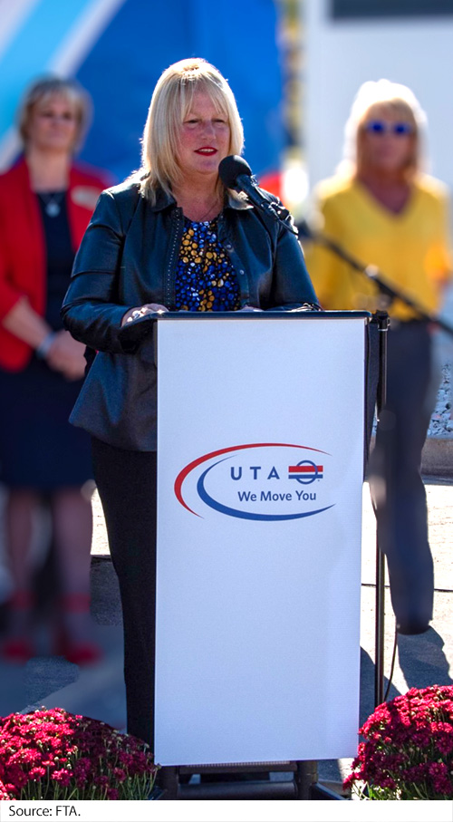 A woman stands at a podium draped with a sign with “UTA, We move you,” with other individuals and a vehicle in the background. Image Source: FTA.