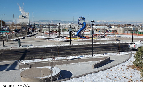 A view from 10 feet above the ground of the Interstate 70 park with a playground, roadway, and concrete sidewalks and walkways, and the Rocky Mountains of Colorado in the far background. Image Source: FHWA.