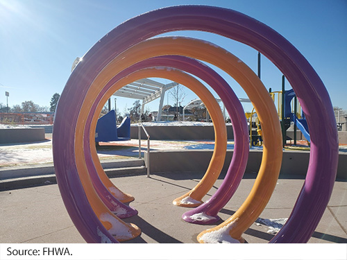 A piece of playground equipment made of several large steel rings standing 6 feet tall on pavement amongst other playground items. Image Source: FHWA.