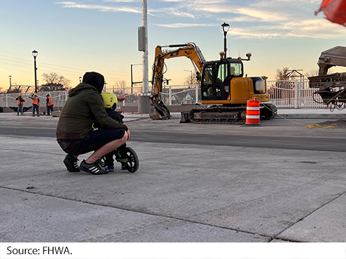 Young child on a small bike, leaning against a man squatting down next to him, looking across the street at construction equipment and workers. Image Source: FHWA.