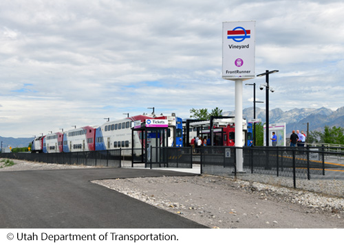 A view of a transit station with trains and people under a cloudy sky and mountains on the right side. Image: © Utah Department of Transportation.