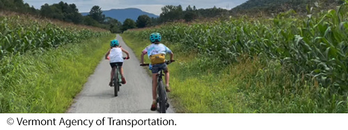 : Two children, each wearing a helmet and riding a bicycle, on a pathway aligned with grass and vegetation. Image: © Vermont Agency of Transportation.